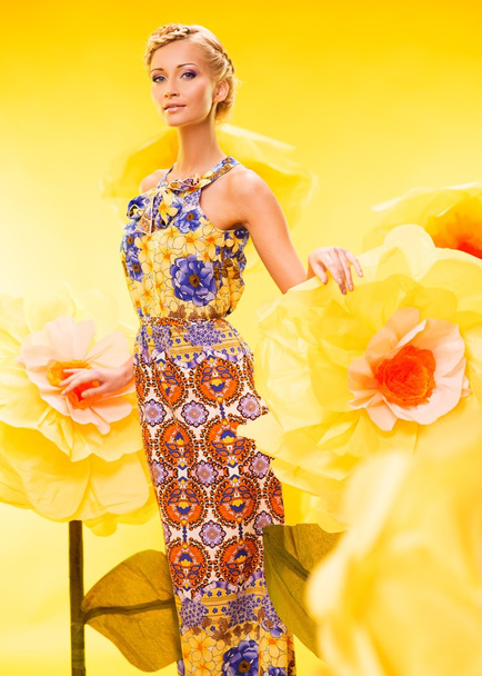Kaunis nuori vaalea nainen värikkäässä mekossa isojen keltaisten kukkien joukossa
 - Valokuva, kuva