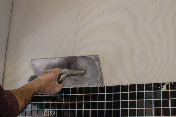 Reparación del hogar: instalación del mosaico en la pared. Un trabajador cubre la pared con adhesivo usando una llana con muescas
. - Foto, imagen