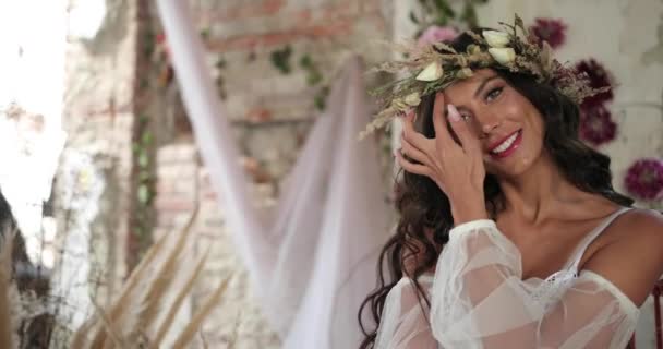 Conceptual shooting. 4k. Woman with long dark hair dressed like a nymph sits on the old bed among blooming pink flowers and hau and smiles looking straight in the camera - Filmagem, Vídeo