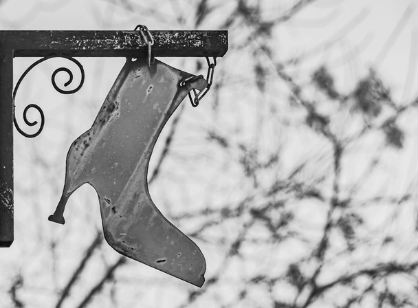 Een teken in de vorm van een oude IJzeren vrouwelijke laars met een scherpe hak op een vage witte achtergrond met takken van een boom kan worden gebruikt voor een schoen reparatiewerkplaats. Zwart-wit foto - Foto, afbeelding