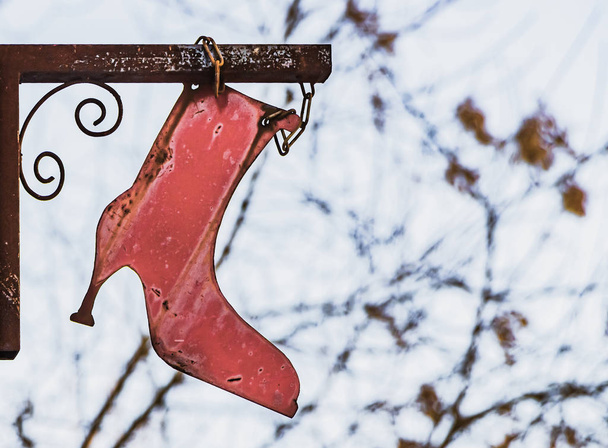 Een teken in de vorm van een oude IJzeren rood vrouwelijke boot met een scherpe hak op een vage witte achtergrond met takken van een boom kan worden gebruikt voor een schoen reparatie fabriek. - Foto, afbeelding