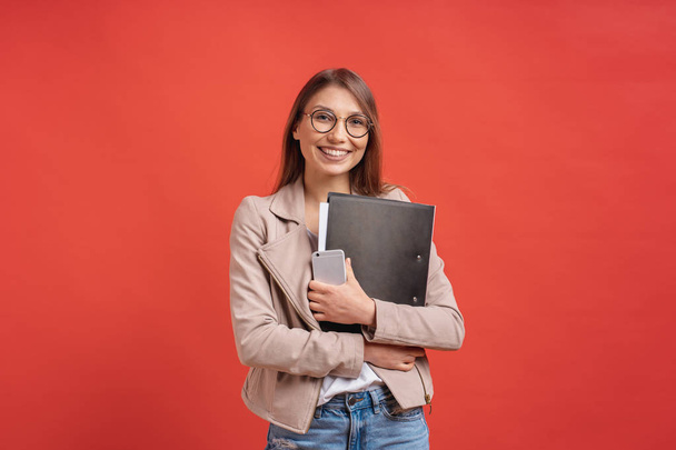 Young smiling student or intern in eyeglasses standing with a folder in hands and happily looking to camera over red background in the studio. - Fotografie, Obrázek