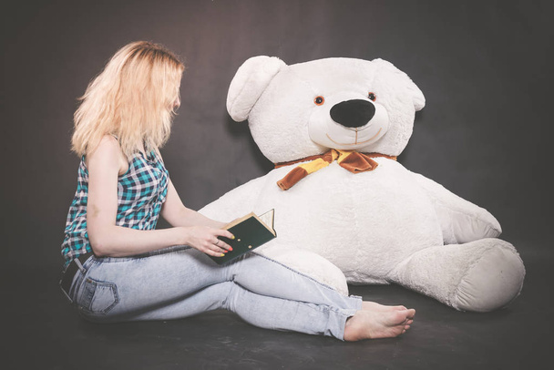cute blonde teen in jeans and plaid shirt plays with her huge Teddy polar bear in yellow scarf on black background alone - Photo, Image