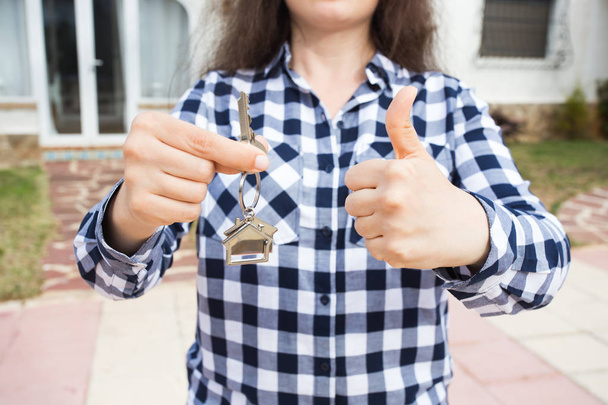 Close up of woman holding key with a keychain in the shape of the house and showing thumbs up. House key on background of new home - Photo, Image