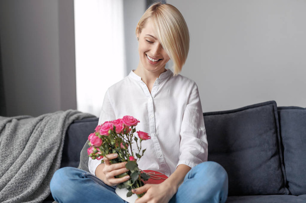 Smiling female with bouquet and gift on sofa - Zdjęcie, obraz