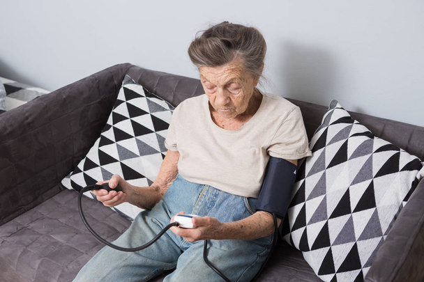 Ця тема є дуже стара людина і здоров'я проблеми. Старший кавказьких жінки, 90-літньому, зморшки і сиве волосся, Головна сидить на дивані і використовує Монітору артеріального тиску для вимірювання кров'яного тиску. - Фото, зображення
