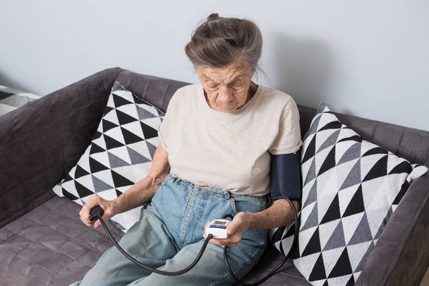The topic is very old person and health problems. A senior Caucasian woman, 90 years old, with wrinkles and gray hair, sits home on sofa and uses blood pressure monitor to measure blood pressure. - Photo, Image