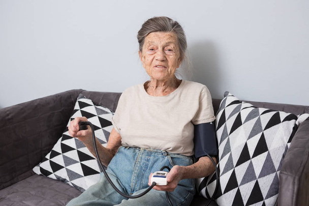Ця тема є дуже стара людина і здоров'я проблеми. Старший кавказьких жінки, 90-літньому, зморшки і сиве волосся, Головна сидить на дивані і використовує Монітору артеріального тиску для вимірювання кров'яного тиску. - Фото, зображення