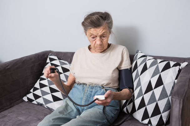 Téma je velmi staré osoby a zdravotní problémy. Vedoucí běloška, 90 let, vrásky a šedivé vlasy, sedí doma na gauči a používá monitor krevního tlaku pro měření krevního tlaku. - Fotografie, Obrázek