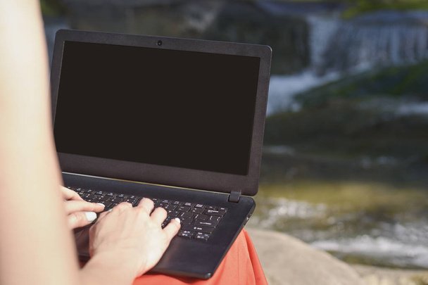 Kobiecych rąk i laptopa z ekranu otwórz na kolanach na tle wodospadu i rzeki. Koncepcja freelance. Pracujesz w przyrodzie. - Zdjęcie, obraz