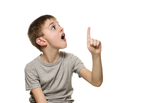 Überraschter blonder Junge in grauem T-Shirt steht auf und zeigt mit erhobenem Zeigefinger. isolieren auf weißem Hintergrund - Foto, Bild