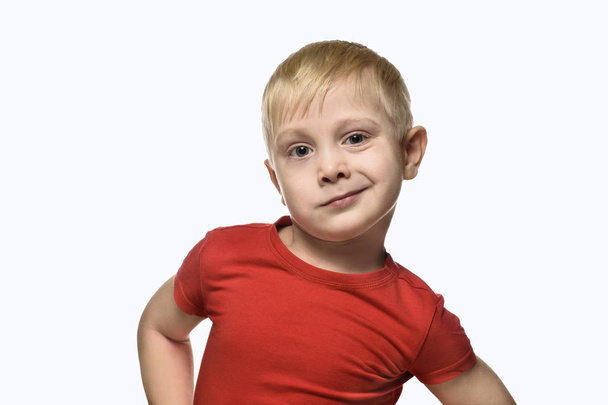 Blonder Junge im roten Hemd, die Arme hinter dem Rücken verschränkt. isolieren auf weißem Hintergrund - Foto, Bild