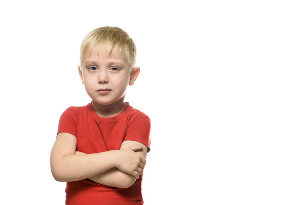 Μικρό ξανθό αγόρι σε ένα κόκκινο πουκάμισο στέκεται squinting με διπλωμένα χέρια. Απομονωθεί σε λευκό φόντο. - Φωτογραφία, εικόνα