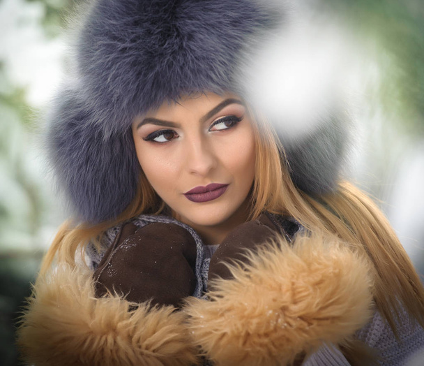 Attractive woman with brown fur cap and jacket enjoying the winter. Side view of fashionable blonde girl posing against snow covered bridge. Beautiful young female with cold weather outfit - Photo, Image