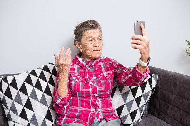 Очень старая кавказская бабушка с седыми волосами и глубокими морщинами сидит дома на диване в джинсах и красной клетчатой рубашке и использует свой смартфон на вытянутой передней камере для видеозвонка
. - Фото, изображение
