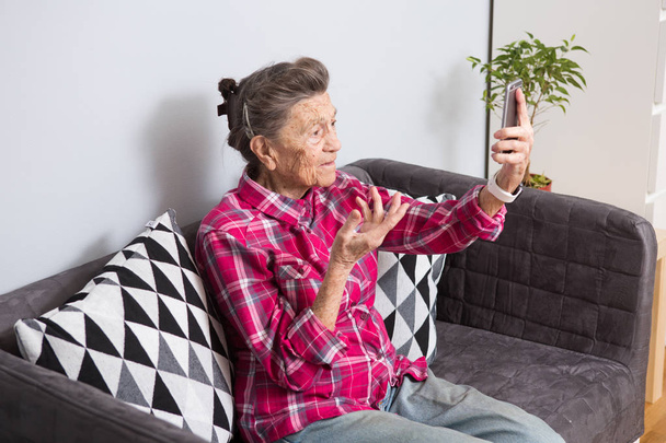 A very old senior Caucasian grandmother with gray hair and deep wrinkles sits home on couch in jeans and red checkered shirt and uses her smart phone on outstretched arm front camera for a video call. - Foto, immagini