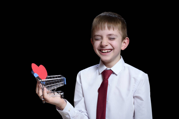 Lächelnder Junge mit geschlossenen Augen hält einen metallenen Einkaufswagen mit einer herzförmigen Postkarte in der Hand. isolieren auf schwarzem Hintergrund - Foto, Bild