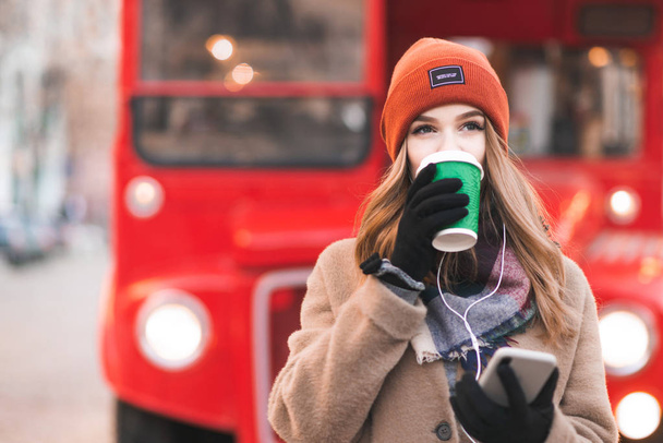 Young lady in a coat and headphones is standing on a red city background with a smartphone in her hands, drinking coffee from a green cup and looking away. Copyspace to the left. - Photo, Image