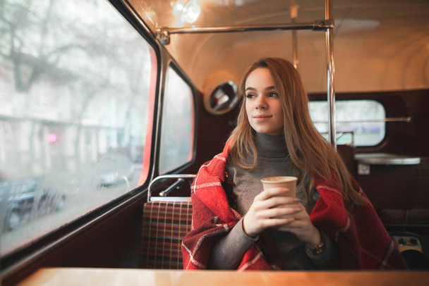 Winter portrait of a girl with a cup of coffee in her hands sits in a coffee shop and warms up covered with a blanket. Attractive girl sits in a retro cafe and looks the window. Cozy portrait. - Фото, зображення