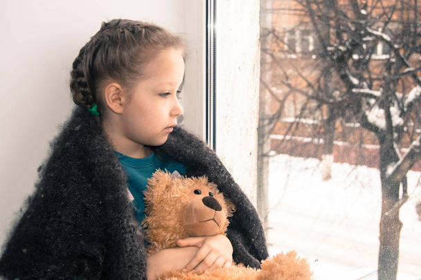 L'émotion du bébé. Une petite fille avec un ours en peluche s'assoit sur le rebord de la fenêtre et regarde par la fenêtre. L'enfant attendait des parents
. - Photo, image