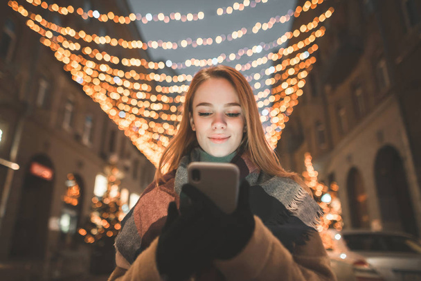 Close-up portrait of a smiling woman uses a smartphone at night on the street street lights. Attractive girl with a smartphone in her hands against the backdrop of an evening night landscape. - 写真・画像