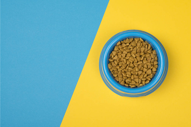 Tigela de alimentação redonda azul com alimentos secos para animais de estimação em um fundo azul e amarelo com espaço para cópia
 - Foto, Imagem