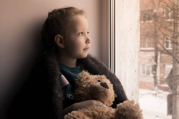 Emoção de bebé. Menina com um ursinho de pelúcia senta-se no peitoril da janela e olha pela janela. A criança estava esperando os pais
. - Foto, Imagem