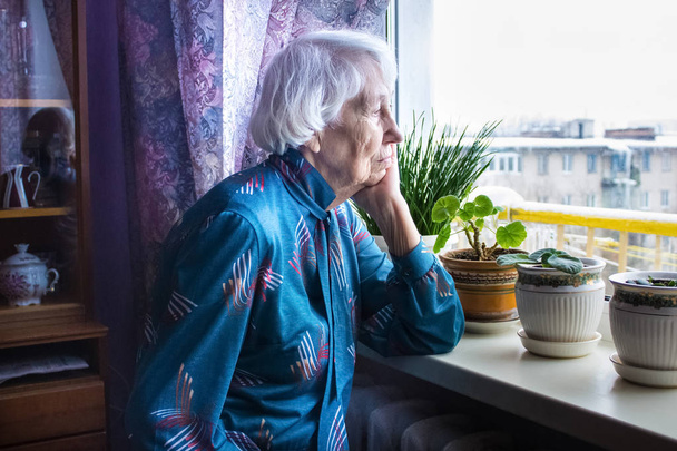 Старая одинокая женщина сидит у окна в его доме. - Фото, изображение