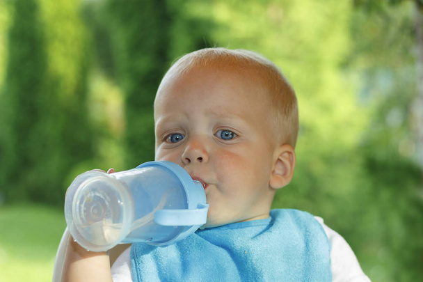 Funny toddler is drinking water from a bottle. It stands on a green lawn - Foto, immagini