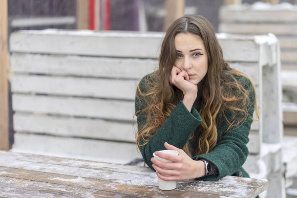 Sad woman with a cup of coffee leaned on a snow-covered wooden table - Foto, Bild