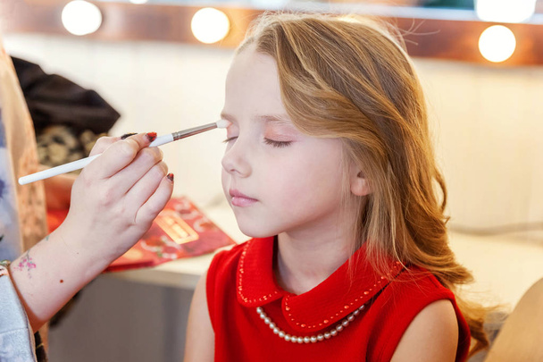 Hand of makeup master applying eyeshadow for little pretty girl model. Professional make up applying by artist. Make-up detail in process. Fashon kid beauty salon photosession concept - Photo, Image