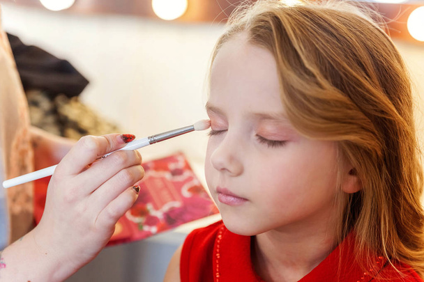 Hand of makeup master applying eyeshadow for little pretty girl model. Professional make up applying by artist. Make-up detail in process. Fashon kid beauty salon photosession concept - Photo, Image