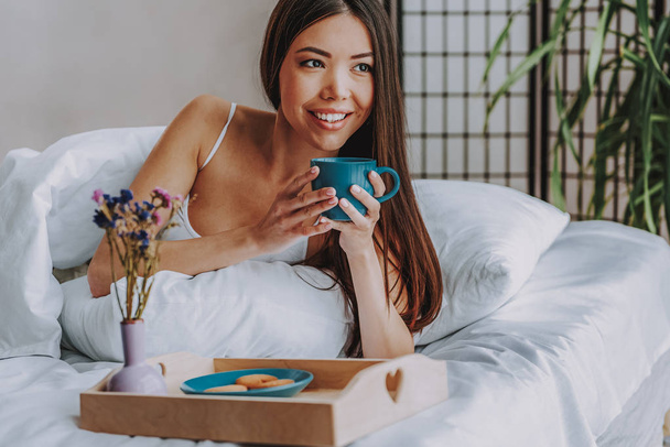 Smiling young female tasting beverage in bed at home - Foto, immagini
