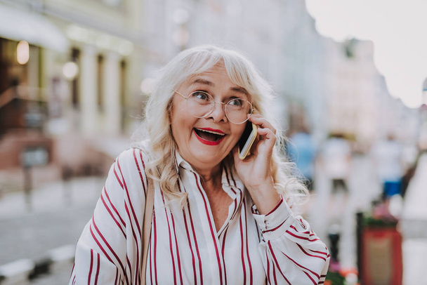 Waist up portrait of old woman. White-haired female with red lips holding hers phone and look happy - Foto, Imagem