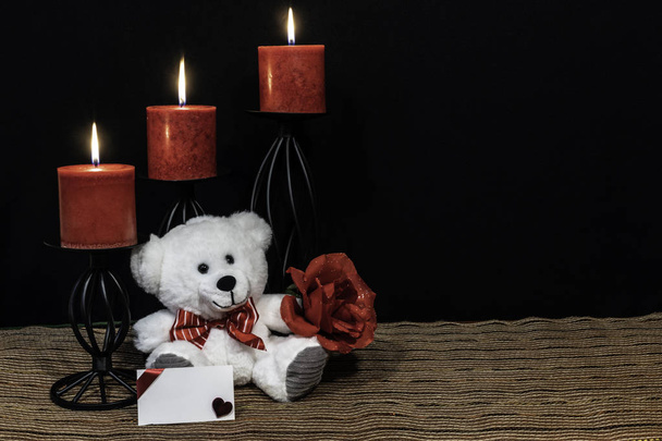 Kuscheliger Teddybär mit roter Fliege, roter Rose, roten Kerzen auf schwarzen Kerzenständern auf Netzmatte und Holztisch mit Karte und dunklem Hintergrund. Valentinstag, Muttertag, Ostern, Weihnachten, Hochzeitskonzepte - Foto, Bild