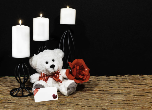 Cudlely teddy bear with red bow tie, red rose, white candles perched on black candle holders on mesh place mat and wooden table with card and dark background. Valentines, Mothers Day, Easter, Christmas, Wedding Concepts - Photo, Image