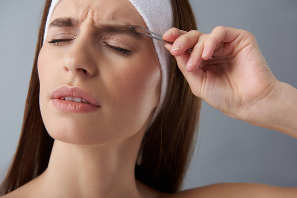 Young woman feeling pain while plucking eyebrows with tweezers - Фото, изображение