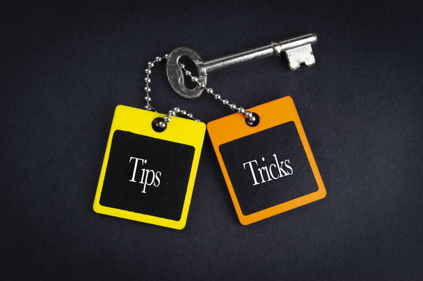 Надпись TIPS TRICKS написана на деревянной бирке и ключе на черном фоне с выборочной фокусировкой и фрагментом урожая. Концепция бизнеса и образования
 - Фото, изображение