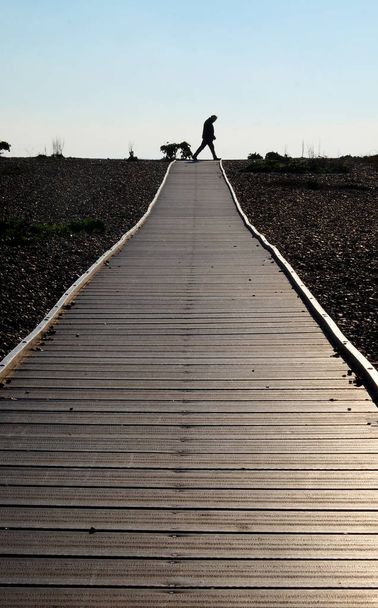 Neerkijkt op een lange rechte houten traject met een kiezelstrand aan beide zijden aan het einde is een onherkenbaar 60-jarige man lopen over afsteekt door de heldere blauwe hemel achter - Foto, afbeelding