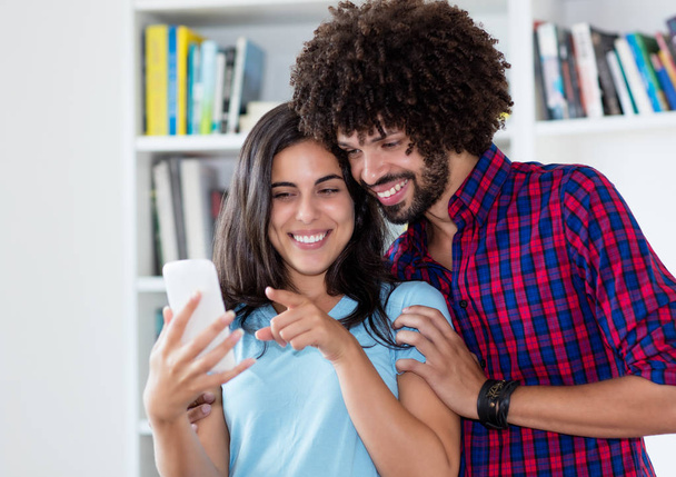 Laughing hipster love couple looking at mobile phone indoors at home - Foto, Imagem