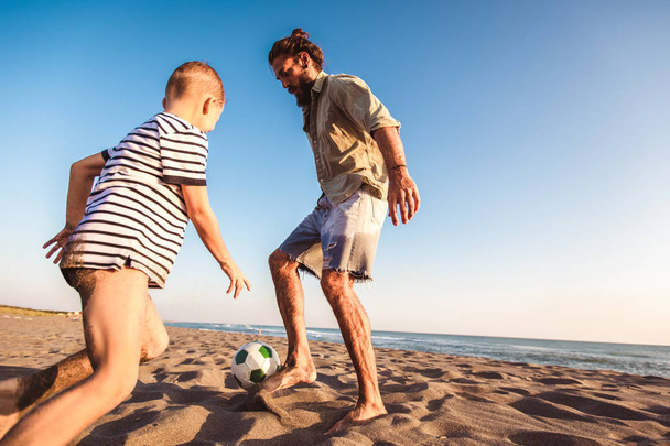 Ευτυχής πατέρας και γιος παίζουν ποδόσφαιρο ή το ποδόσφαιρο στην παραλία έχοντας μεγάλη οικογενειακή στιγμή για τις καλοκαιρινές διακοπές. - Φωτογραφία, εικόνα
