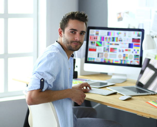 Retrato del joven diseñador sentado en el estudio gráfico frente a la computadora portátil y el ordenador mientras trabaja en línea. - Foto, Imagen