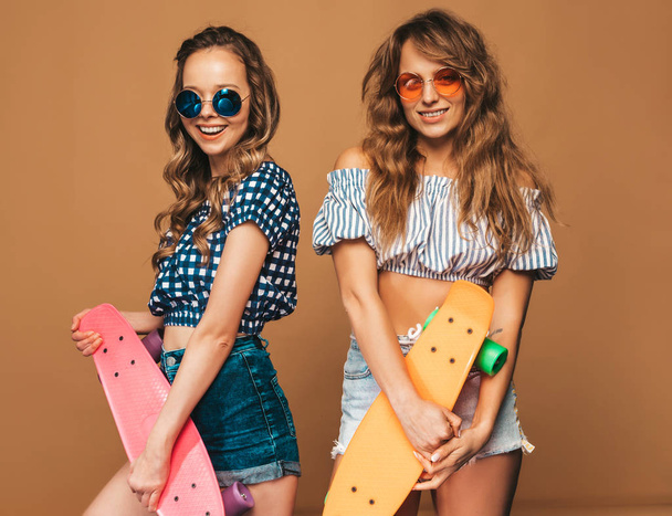 Two young stylish smiling beautiful girls with penny skateboards.Women in summer hipster checkered shirt clothes posing near golden wall in sunglasses. Positive models having fun and going crazy - Fotó, kép