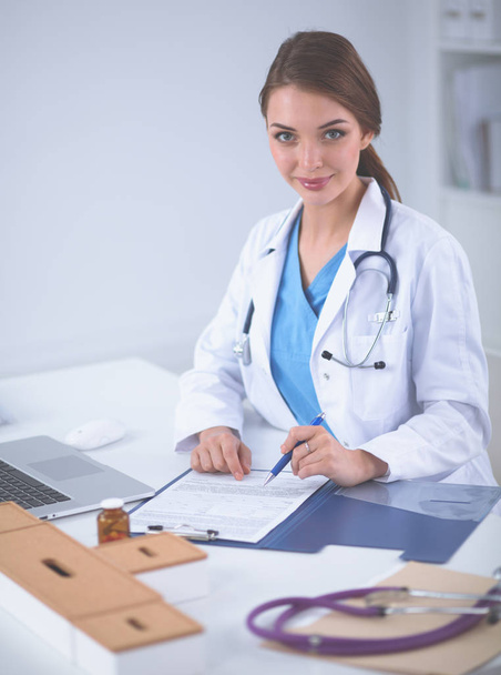 Beautiful young smiling female doctor sitting at the desk and writing - Foto, afbeelding