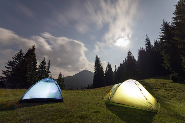 Bright big moon in dark blue cloudy sky over two tourist tents on green grassy forest clearing among tall pine trees on distant mountain background. Tourism, night camping in summer mountains. - Photo, Image