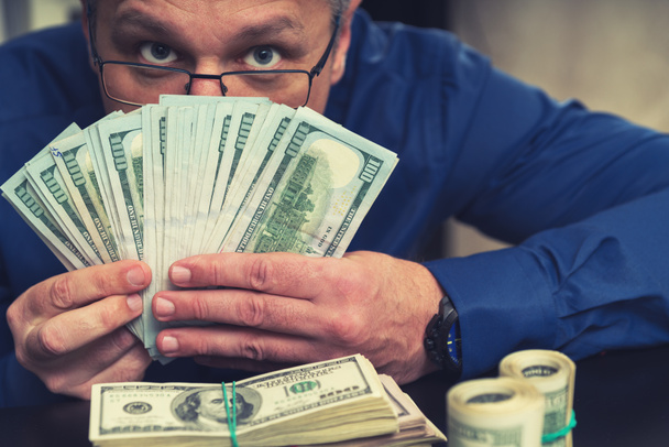 Hombre de negocios subrepticio escondido detrás de un puñado de billetes de dólar mirando por encima de la cámara en un concepto de juego, éxito, negocios, ganancias o soborno o pago
 - Foto, Imagen
