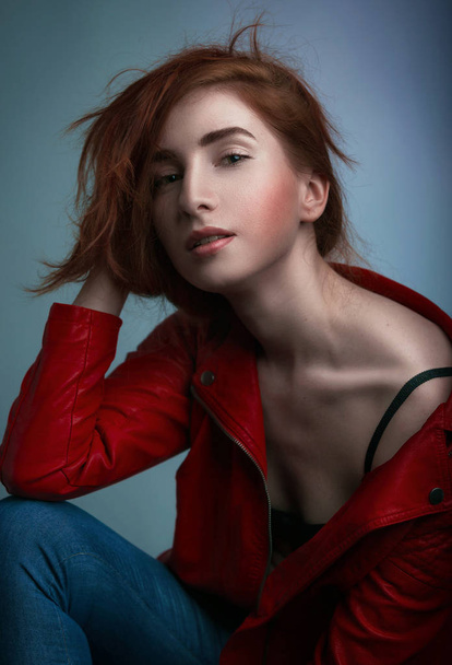 Studiový portrét ženy s červenými dlouhými vlasy červené kožené bundě, otevřené ramenní sedí na židli teplých tónů, módy a stylu koncepce - Fotografie, Obrázek