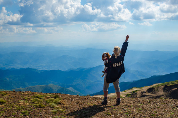 Mujer sosteniendo a su perro con un brazo levantado, en una cima de la montaña. Traducción del texto de la chaqueta: "Serbia
". - Foto, imagen
