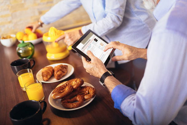 Ouder paar drukke kijken naar digitale tablet terwijl het hebben van heerlijk ontbijt thuis keuken - Foto, afbeelding