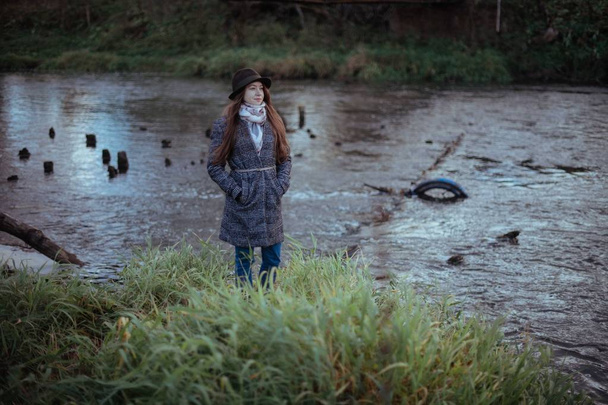 Dziewczyna stoi na brzegu rzeki, w pobliżu mostu na pochmurny wieczór. Młoda kobieta w płaszcz i kapelusz stoi nad wodą - Zdjęcie, obraz